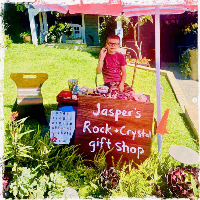Jasper, who is approximately nine years old, stands behind a wooden sign saying "Jasper's Rock + Crystal Gift Shop", with a pink canopy overhead and a board showcasing various crystal and rock pendants beside him.