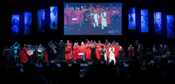 GLIDE musicians onstage singing in red robes with screen overhead simulcasting their performance.