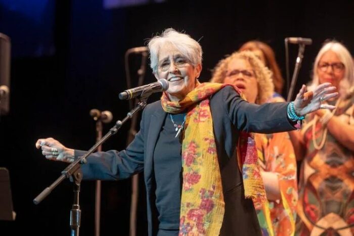 Joan Baez, wearing a gray wool suit and an orange print scarf, smiles as she sings on-mic with backup vocalist in the background.