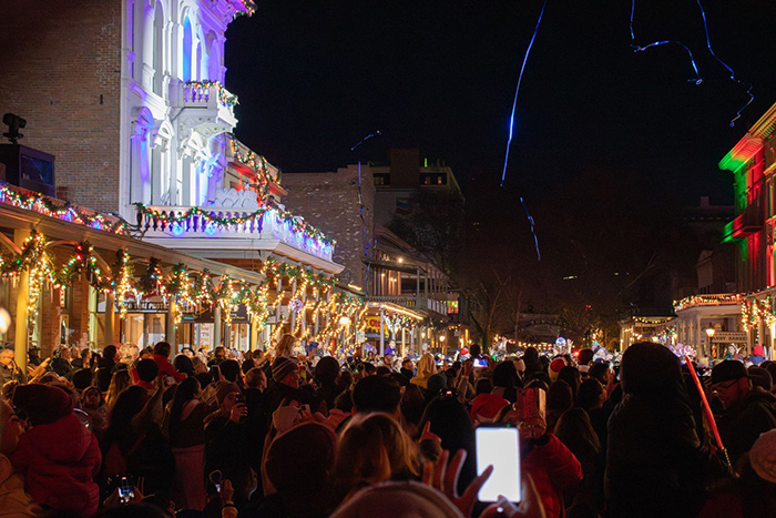 Crowd stands under Old Sacramento wooden balcony illuminated with white lights.