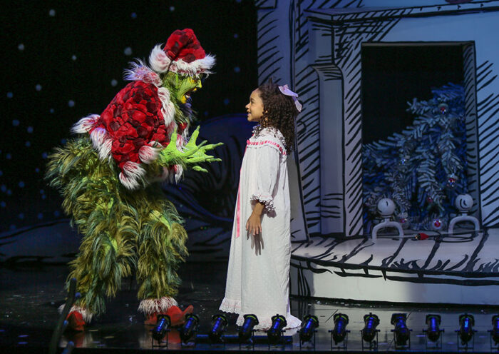 Andrew Polec, as the Grinch dressed in a Santa hat and red jacket , splays his hands and looks startled as Cindy-Lou Who (Arden Johnson), in a white nightgown, asks him what he's doing.