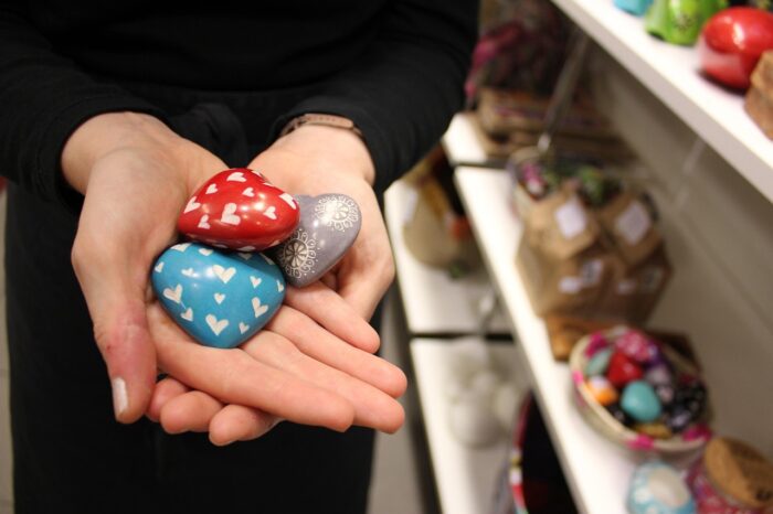 Woman's hands as she holds three painted ceramic hearts: a red, a white and a blue, each panted with smaller hearts, near a set of store shelves with additional merchandise.