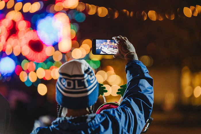 Woman in a blue jacket and a COLTS knitted cap raises her phone to photograph the tree lights, in the distance.