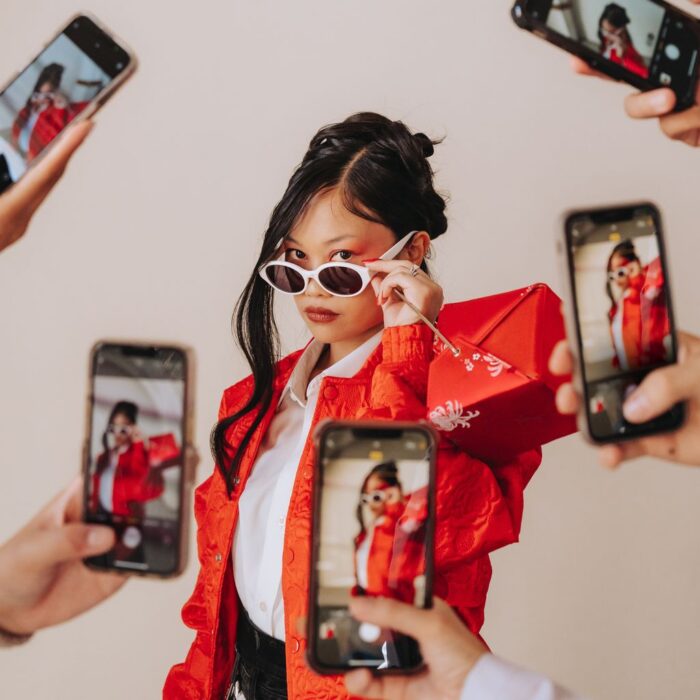 Ruby Ibarra, wearing a red jacket and white shirt, looks over her sunglasses as four camera phones surround her to snap a picture.