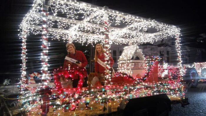 Two women with beauty-queen sashes and red sweaters ride a float lighted with silver lights and with a giant milk carton at the back.