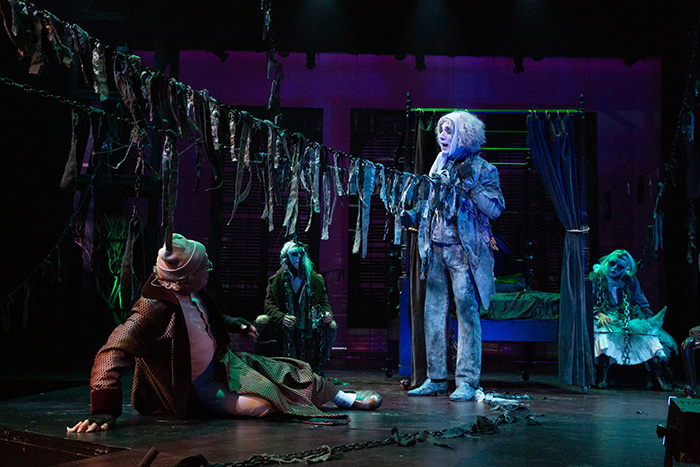 Scrooge sits on floor and looks up in terror at the ghost of Jacob Marley, lit in blue lights.