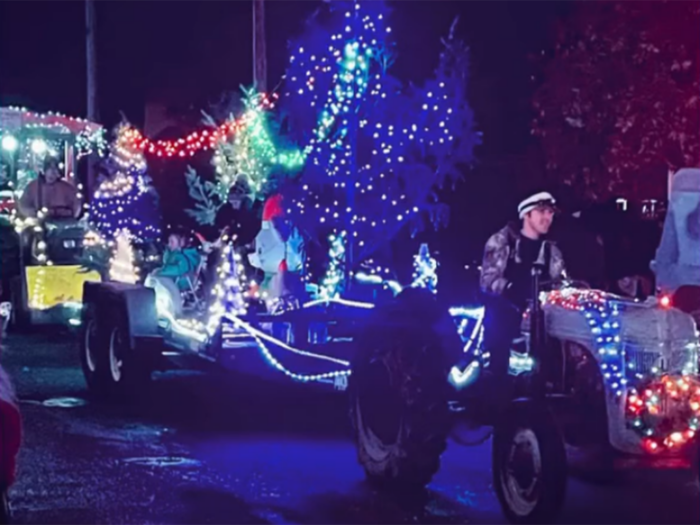 Young man drives a lighted tractor, pulling a float with Christmas trees, a lighted cat and blowing "snow".