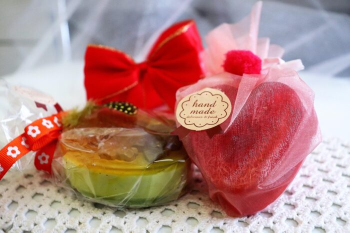 Two handcrafted soaps, one red one in the shape of a heart and one round green one, on a white lace cloth with a red bow on top of them.