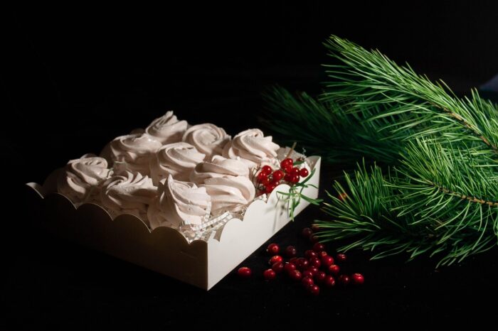 Box of a dozen whipped white meringue cookies with red cinnamon drops next to a Christmas tree branch.