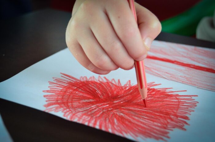 Child's hand holding an orange drawing pencil as they create a starburst design on drawing paper.