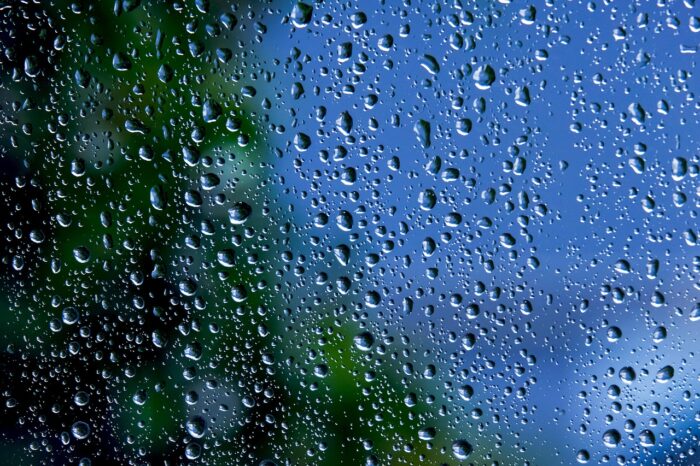 Raindrops on a window with blurred sky and foliage beyond.