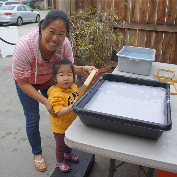 Smiling mom and her small daughter stand near a screenprinting vat as the child holds the print they've just made.
