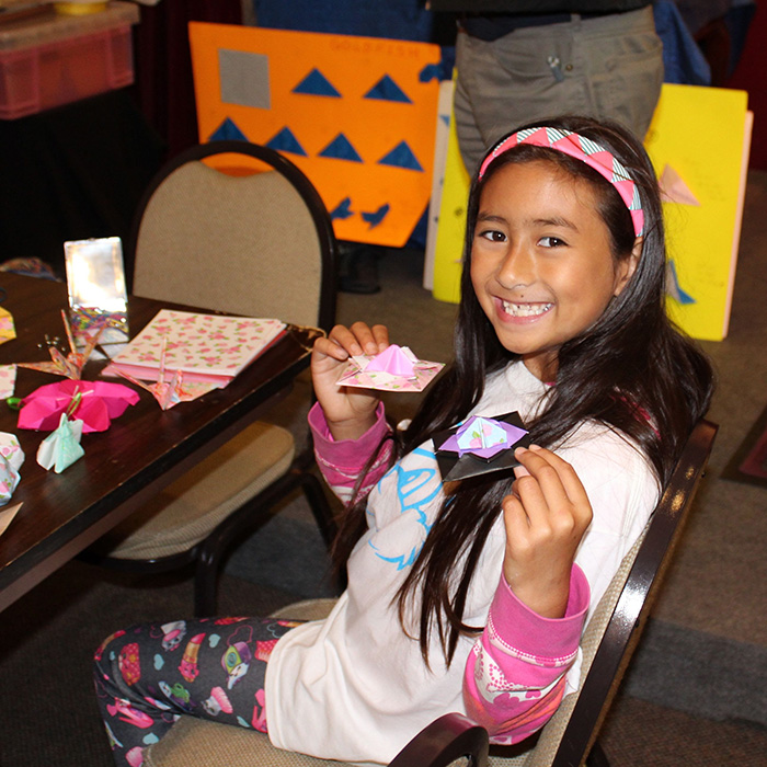 Young girl, about eight years old, holds up the origami paper cranes she made while sitting at a table.