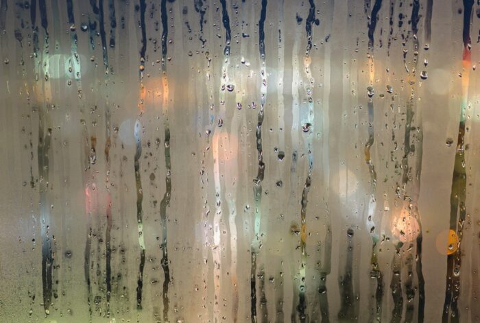 Windowpane with rain, condensation and multicolored lights beyond it.