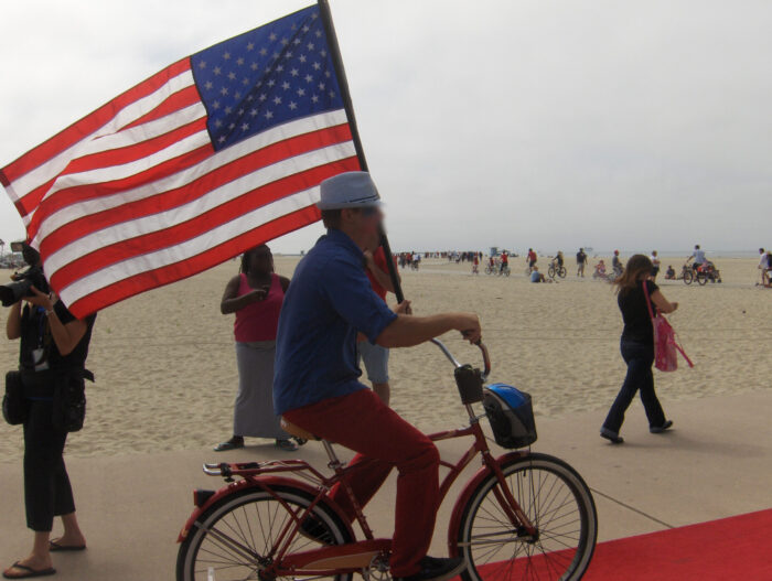 Man holding full-sized American flag as he rides down the red carpet to the bike path on his bicycle.