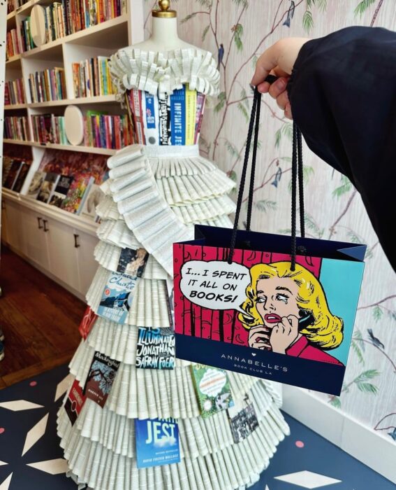 Woman's arm holding a tote bag with cartoon of a woman crying on the phone, "I--I spent it all on BOOKS!", and filled bookshelf in the background