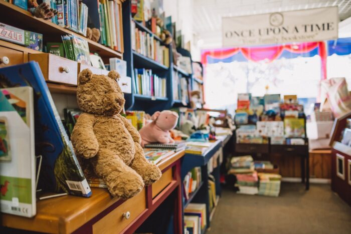 Teddy bear sits on bookshelf near floor-to-ceiling book display with "Once Upon a Time" sign in the distance