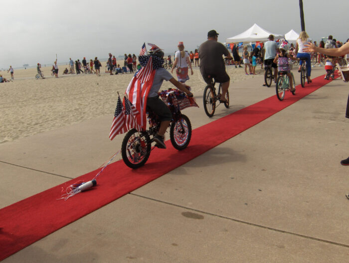 Bystander applauds a young boy riding a bike festooned with flags and pulling a silver "rocket" made of cardboard and tinfoil