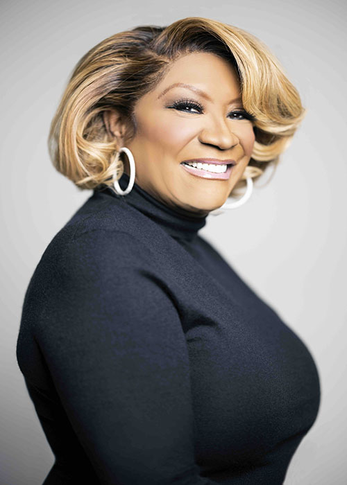 Patti LaBelle, in a black turtleneck and silver hoop earrings, smiles as she stand sideways for a headshot.