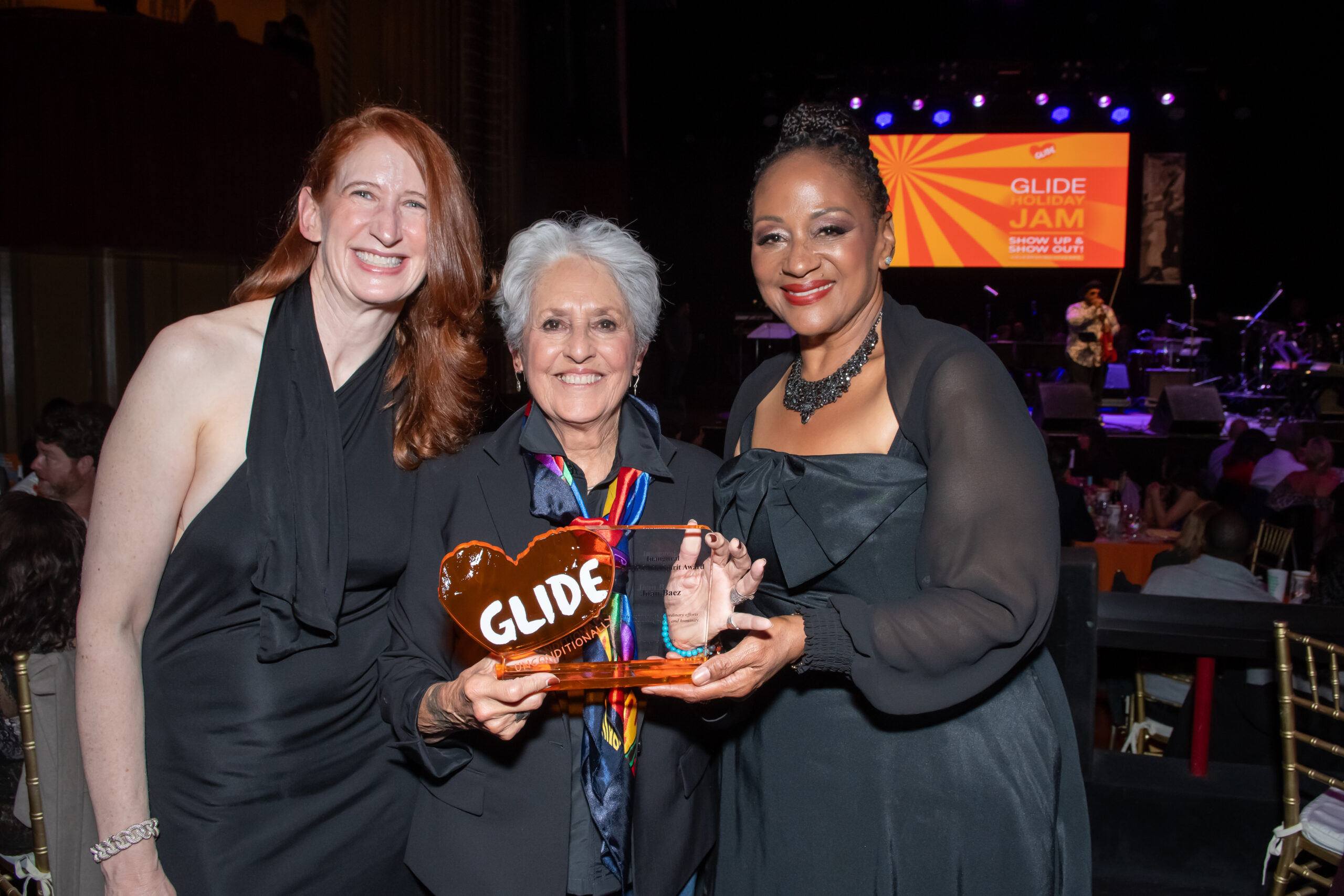 Inaugural Lizzie Glide honoree Joan Baez, flanked by two presenters, smiles as she displays her heart-shaped award onstage.