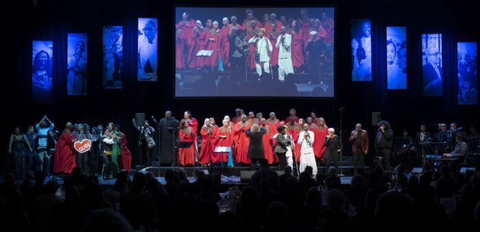 GLIDE Ensemble and The Change Band perform onstage with a giant monitor behind them and director Zoe Ellis leading them.