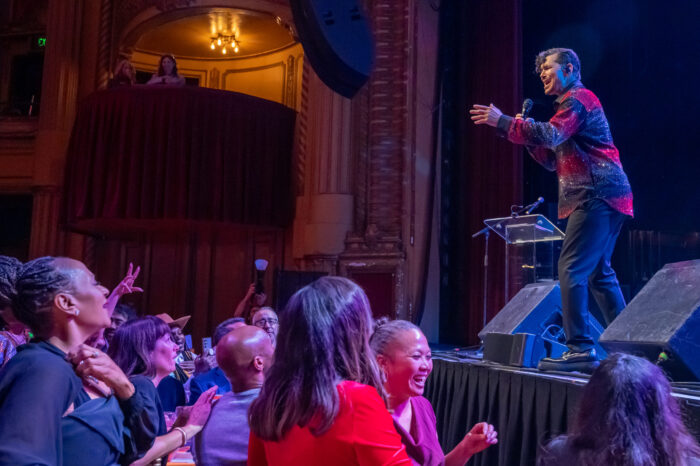 Smiling performer in blue-and-red striped sweater with jeans onstage as audience members smile.